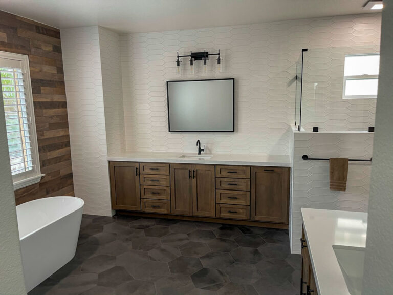 The image shows a simple bathroom vanity with a rectangular ceramic sink. The faucet has two handles and a polished chrome finish. Below the sink are two cabinets with white shaker-style doors and chrome handles. A large rectangular mirror is mounted on the wall above the sink. To the left of the vanity is a white bathtub.