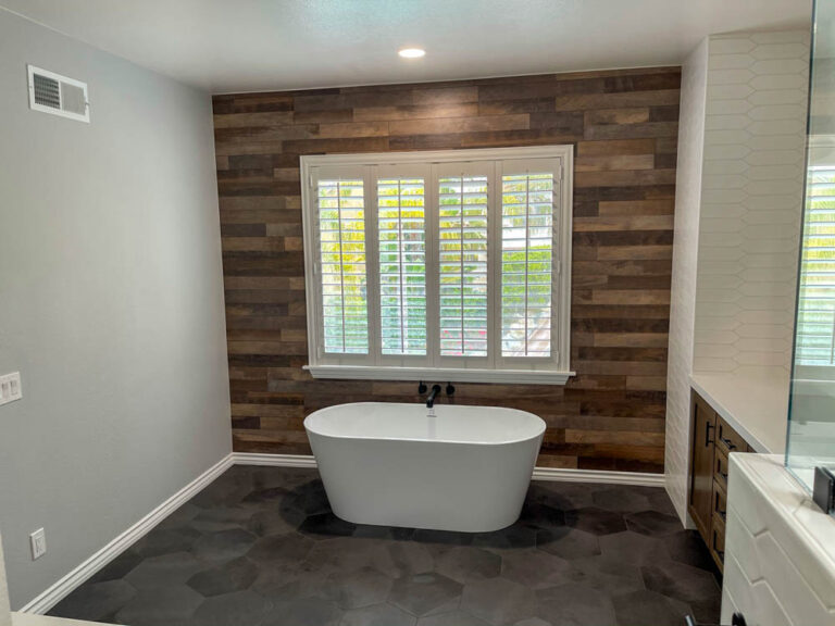 A simple yet inviting bathroom retreat. A white bathtub sits nestled beneath a window, framed by a warm wooden wall. Sunlight streams through the window, casting a soft glow on the clean lines of the tub. The uncluttered space and natural light create a sense of peace and tranquility, perfect for a relaxing soak at the end of a long day.