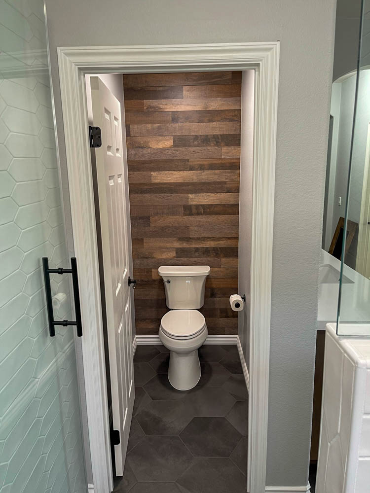 A toilet area with black tile flooring. A tissue holder mounted on the wall. And the toilet wall is made of hardwood floor giving a rustic accent.