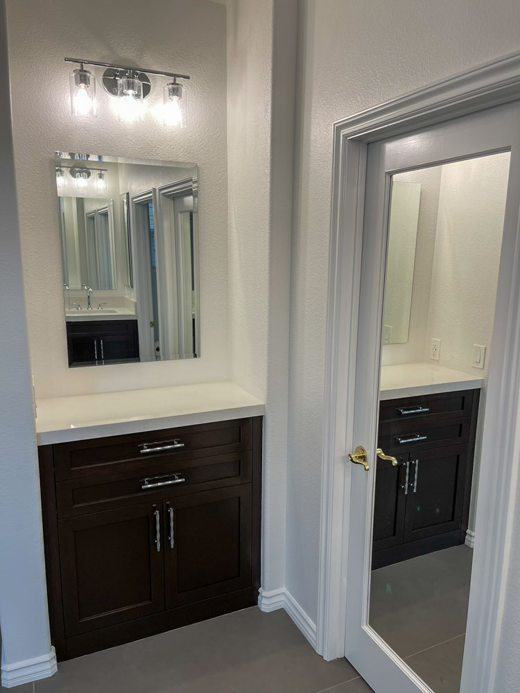 A vanity area in a bathroom. There is a wood cabinet with a ceramic countertop and a mirror just above. There is a wall-mounted lighting fixture for ample illumination.