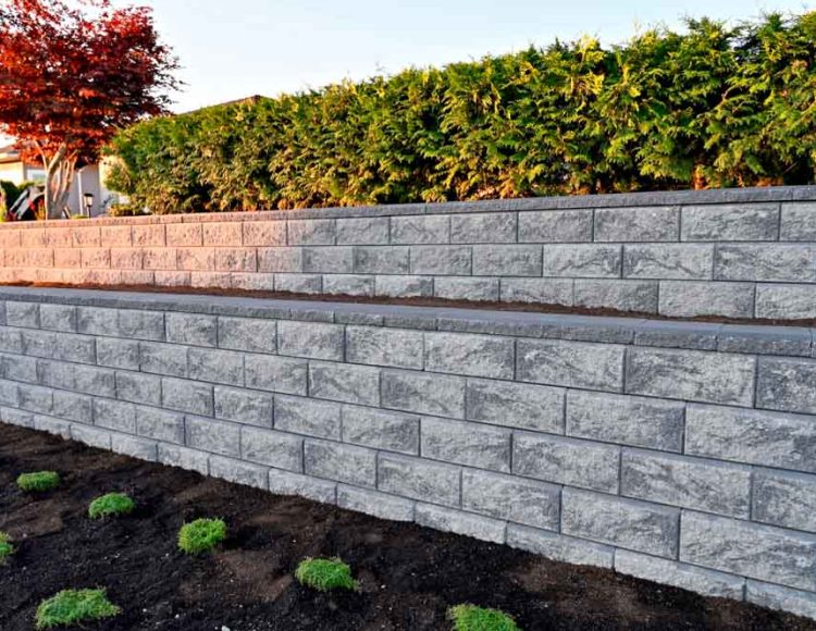 A newly constructed retaining wall made of textured gray blocks in a residential garden during the golden hour, casting soft light across the scene. The wall is tiered, with small green plants at its base and a lush hedge behind it. The environment suggests a well-maintained outdoor area designed to enhance the landscape and provide soil support.
