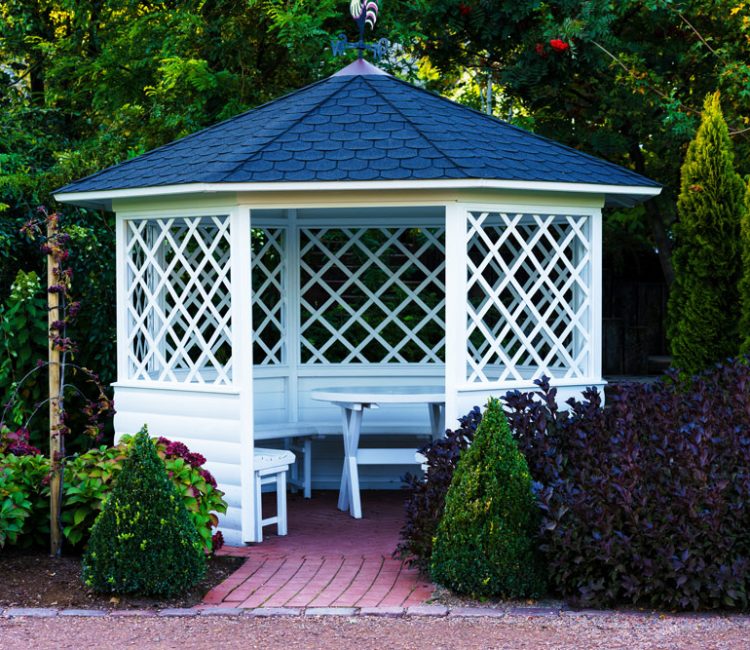 A charming white gazebo with a bright blue roof nestled amidst a lush green garden. The gazebo appears to be constructed from a latticework material, allowing light to filter through and creating a sense of airiness. The roof of the gazebo is a vibrant blue color, creating a delightful contrast with the white latticework and the surrounding greenery. A walkway, paved with light-colored stones, leads up to the entrance of the gazebo. Surrounding the gazebo on all sides are a variety of plants and flowers. Overall, the image conveys a feeling of peace and tranquility, making the gazebo seem like a perfect spot for relaxation and reflection in a beautiful garden setting.