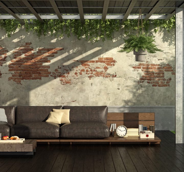 A stylish outdoor lounge area with a dark leather sofa against a rustic, exposed brick wall partially covered in peeling plaster. Overhead, a pergola with lush greenery provides shade, casting shadows on the wall. The space is bordered by clear glass panels, offering views of a well-manicured garden and a tree with ripe oranges. A vintage bicycle and modern low-profile coffee table complete the serene, inviting ambiance.