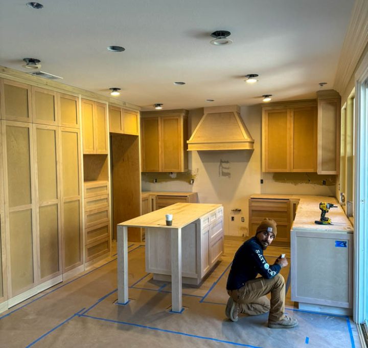 A modern kitchen under renovation. A man, wearing casual attire with a bonnet hat, is sitting and smiling at the camera. The room features unfinished wooden cabinetry and a central island, both complemented by a sleek design. The kitchen's floor is covered with protective blue tape, indicating ongoing work. Recessed lighting is installed in the ceiling, enhancing the contemporary feel of the space.