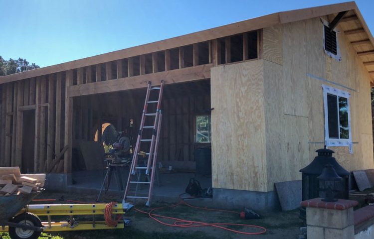 Construction of a wooden structure with a partially completed plywood and timber framework under a clear blue sky. A worker can be seen in the open garage area, surrounded by various construction tools, including a ladder, a saw, and electrical cords. The building features exposed beams and studs, and a new window installed in the side wall, indicating ongoing work in a residential project.