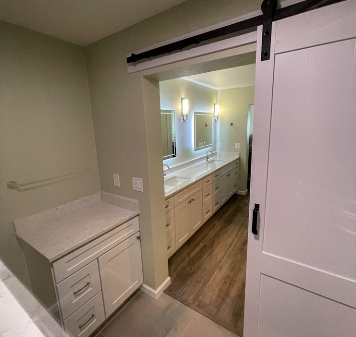 A shower area with tiled flooring and a barn door for partition with the vanity area. A storage solution on one corner, and a towel bar just above.