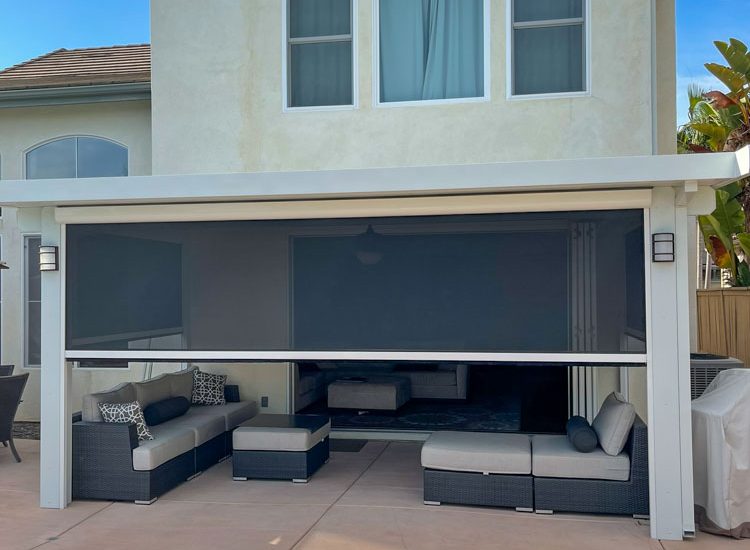 Exterior view of a two-story home with an extended patio cover featuring large retractable shades, providing flexible sun protection. The patio is equipped with modern black wicker furniture, including sofas and ottomans adorned with stylish blue and white cushions. This setup overlooks a residential backyard with a glimpse of a pool, reflecting a functional and attractive outdoor living space ideal for relaxation and entertainment. The house exterior is finished in a soft beige stucco, complemented by a clear blue sky.
