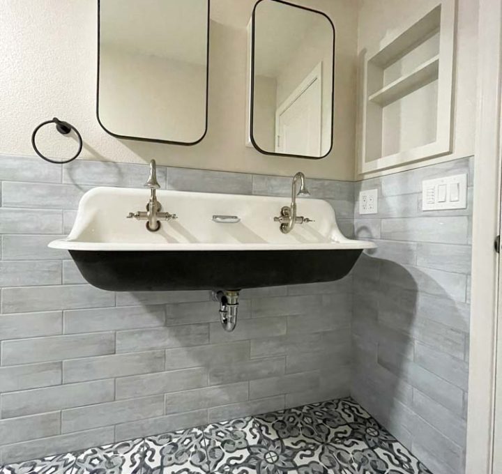 A stylish bathroom featuring a large industrial-style wall-mounted sink with dual faucets set against a grey tiled backdrop. Above the sink, two rectangular mirrors are framed by sleek black fixtures. The floor is adorned with ornate black and white tiles that add a decorative touch to the functional space. The design combines modern and industrial elements, creating a clean and sophisticated atmosphere.
