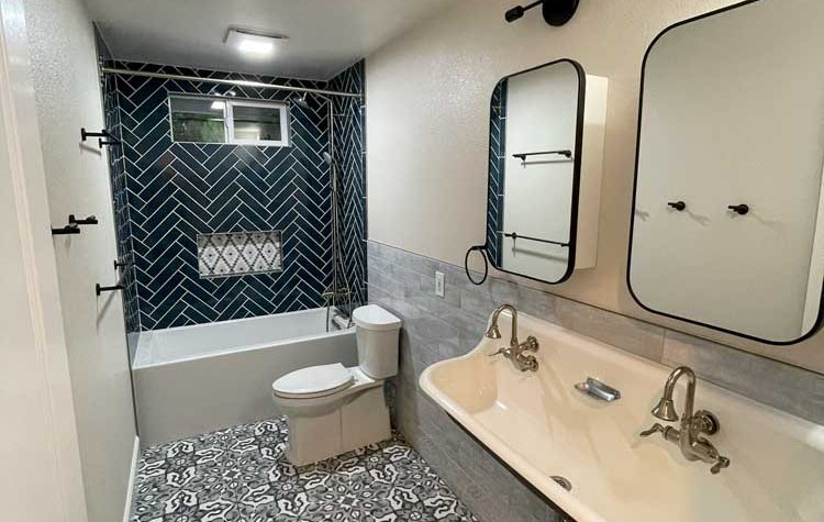 A beautifully designed bathroom featuring a mix of modern and vintage elements. The room is equipped with a large bathtub with a unique navy blue and white herringbone tile surround and a glass shower partition. Two rectangular mirrors with rounded edges and black frames are mounted above a long, classic white sink with vintage-style faucets. The floor is covered in ornate black and white tiles that add a dramatic flair. This space blends contemporary design with traditional accents, creating a striking visual impact.