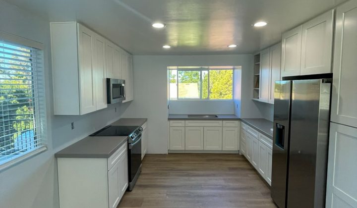 A modern kitchen with clean lines and a minimalist aesthetic. The kitchen features white cabinets with stainless steel pulls and countertops. There is a stainless steel refrigerator and a dishwasher. The kitchen is well-lit with recessed lighting.