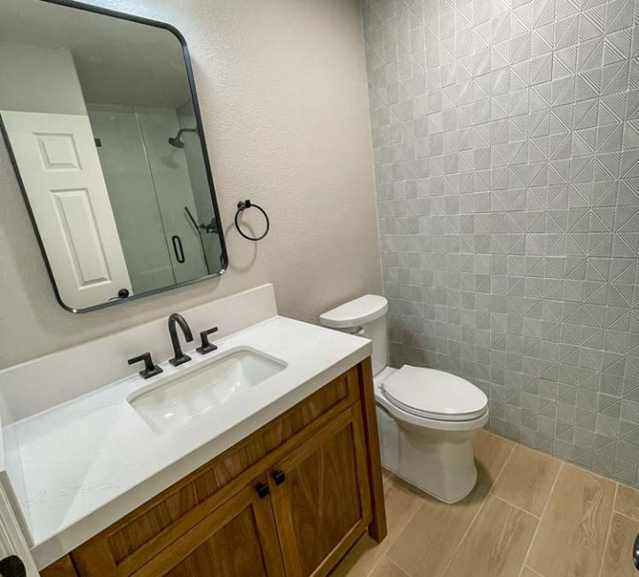 A modernized bathroom with neutral gray walls, geometric patterned tiles, a dark wood vanity with a white countertop, a stylish rectangular mirror, and updated black fixtures.