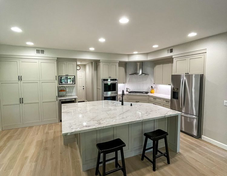 A spacious and modern kitchen illuminated by recessed ceiling lights, featuring pale grey cabinetry and a large central island with a marble countertop. The island includes a sink and is accompanied by two black bar stools, providing a casual dining area. Stainless steel appliances, including a refrigerator, oven, and microwave, complement the sleek design. The kitchen's light wood flooring adds warmth to the overall minimalist aesthetic.